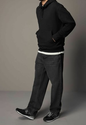 Person wearing black hoodie, white undershirt, dark gray jeans, and black sneakers standing against a plain neutral background.