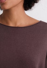 Brūns adīts tops ar plašu, izgrieztu apkakli. Audumam ir gluda tekstūra, ar smalku ribotu detaļu ap apakšmalu un apkakli.