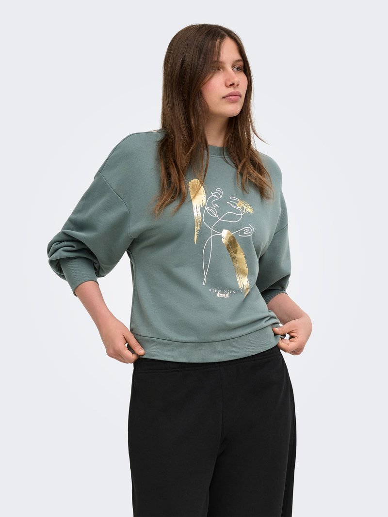 Jeune femme aux longs cheveux bruns portant un sweat-shirt vert avec un motif abstrait doré et un pantalon noir, posant en ajustant l'ourlet avec les mains.
