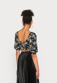 Blusa curta preta com padrão floral, mangas bufantes e decote profundo, combinada com uma saia preta plissada de cintura alta.