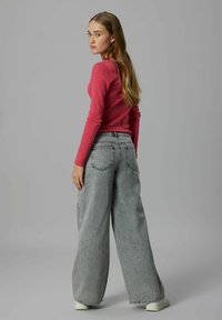 Ung kvinne med langt hår iført en rosa genser med lange ermer, grå vide jeans og hvite joggesko, stående mot en grå bakgrunn.