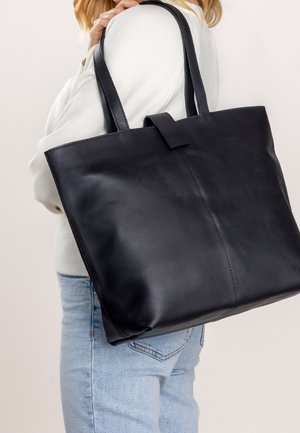 Femme portant un pull blanc et un jean clair, tenant un grand sac fourre-tout en cuir noir sur l'épaule, sur un fond uni.