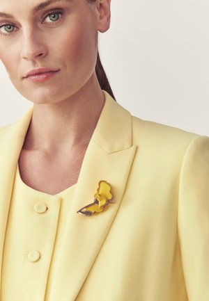 Femeie purtând un sacou și un top galben deschis, cu o broșă galbenă în formă de floare, asortată, prinsă pe reverul sacoului.