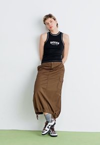 Svart tanktopp med "DICKIES"-logo, brun cargo-skjørt med innsnørt hem, grå sokker og svart-hvite joggesko. Glatt stoff, avslappet passform.