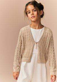 Cardigan crema lavorato a maglia con motivo in pizzo aperto, maniche corte e chiusura a fiocco al collo. Indossato sopra un vestito bianco, presenta polsini texturizzati.