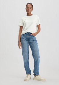 Witte t-shirt met ronde hals, gecombineerd met hoge taille blauwe jeans. Het model draagt lichte sneakers en heeft een natuurlijke haardos.