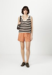 Ραβδωτό πλεκτό αμάνικο μπλουζάκι σε μαύρο και κρεμ, συνδυασμένο με πορτοκαλί σορτσάκι. Το μοντέλο κρατάει μια μικρή καφέ τσάντα, φορώντας σκούρες παντόφλες.