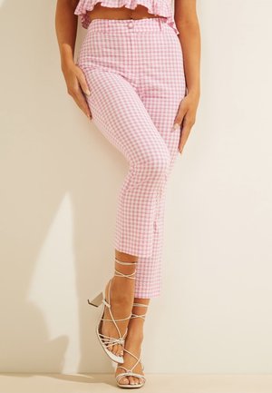Femme portant un pantalon vichy rose taille haute et des sandales blanches à talons avec des brides, debout contre un mur beige uni.
