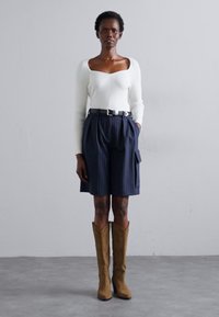 Hvit ribbet langermet topp, marineblå rutete skreddersydde shorts med sidelomme, og brune cowboyboots med dekorativ stikning.