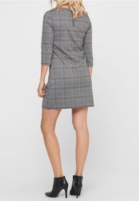 Robe à carreaux grise avec des manches trois-quarts, dotée d'une fermeture éclair dans le dos. Portée avec des bottines noires, présentée de dos.