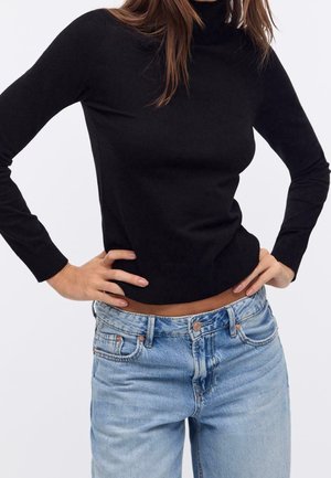 Femme portant un pull à col roulé noir ajusté à manches longues et un jean taille haute bleu clair, les mains sur les hanches.
