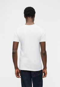 Άνδρας με την πλάτη στραμμένη, φοράει λευκό απλό κοντομάνικο μπλουζάκι και σκούρο τζιν με κόκκινο κεντητό σχέδιο πουλιού στην πίσω τσέπη.