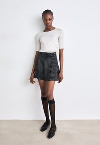 Lichtgrijze aangesloten top in combinatie met een zwarte geplisseerde rok met witte verticale strepen. Zwarte kniehoge sokken en zwarte ballerina's maken de look compleet.