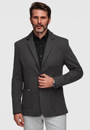 Uomo con barba corta che indossa una giacca grigio scuro sopra una camicia nera, sta allacciando un bottone con la mano destra, pantaloni grigio chiaro, orologio d’argento al polso sinistro.