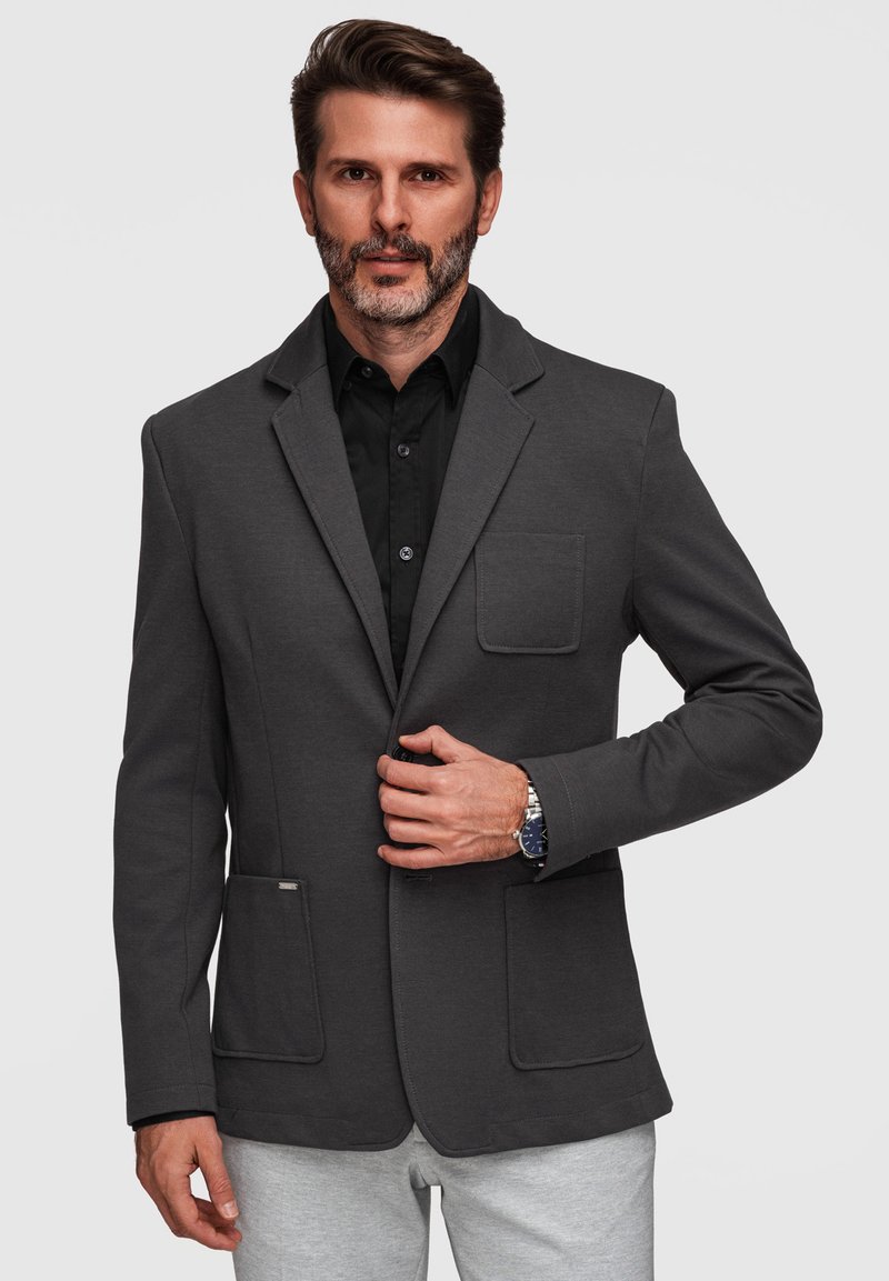 Homme avec une barbe courte portant un blazer gris foncé sur une chemise noire, boutonnant avec la main droite, pantalon gris clair, montre argentée au poignet gauche.