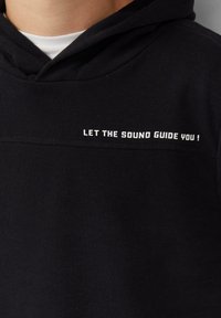 Zwarte hoodie met witte tekst "LAAT DE GELUIDEN JE GIDS ZIJN !" gedrukt op de borst, gedragen over een wit onderhemd.