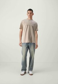 Jovem vestido com uma t-shirt bege com logo, calças de ganga largas desbotadas e ténis brancos, de pé contra um fundo claro e simples.