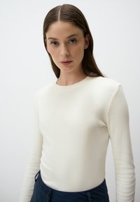 Top bianco a maniche lunghe con scollo rotondo, silhouette aderente e texture liscia, abbinato a dettagli in vita blu navy.