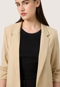 Bežový blazer se střižením na míru a měkkou tkaninou, s vystuženými rameny a ohrnutými rukávy, kombinovaný s černým ribkovaným topem.