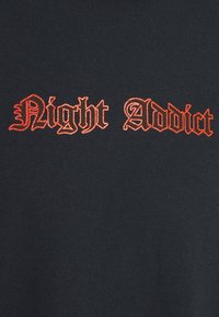 Svart tyg med texten "Night Addict" i en fet, gotisk stil broderad i orange, centrerad på plagget.