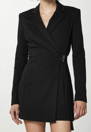 Frau trägt ein schwarzes Wickelblazerkleid mit langen Ärmeln und seitlichem Schnallenverschluss, steht vor einem schlichten hellen Hintergrund.
