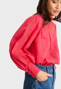 Blusa de algodón roja con cuello alto, detalles bordados y mangas voluminosas. Los puños abotonados y los paneles texturizados añaden interés visual.