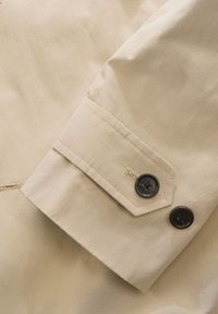 Beige overhemdmanchet met twee zwarte knopen en een diagonale naad. De stof heeft een gladde textuur met minimale kreukels.