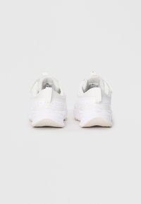 Une paire de baskets de sport blanches vues de l'arrière sur un fond gris clair uni, avec des tirants et des semelles texturées.