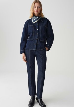 Dunkelblaue Jeansjacke mit zwei vorderen Taschen und silbernen Knöpfen, kombiniert mit dunkelblauen Jeans im geraden Schnitt. Schwarze Stiefel runden den Look ab.