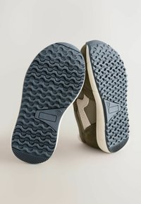 Trampki mają teksturowaną granatową gumową podeszwę, oliwkową tkaninę na cholewce i beżowe akcenty. Design zawiera wyraźny bieżnik dla lepszej przyczepności.