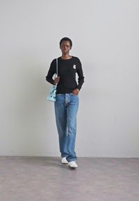 En person går självsäkert, iklädd en svart tröja med en knapp, en ljusblå väska, lösa jeans och vita skor, mot en ljusgrå bakgrund.