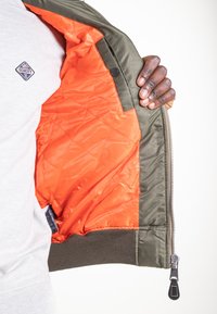 Giubbotto bomber verde oliva con zip aperta che mostra l'imbottitura trapuntata arancione brillante. Presenta polsini a coste e chiusura con zip con cursore in pelle.