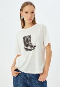 Hvid bomulds t-shirt med rullede ærmer, der viser en grafik af sorte cowboy støvler med pink accenter og blomsterdesigns.