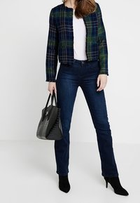 Veste en tweed à carreaux bleu foncé, vert et noir, associée à un jean denim foncé ajusté. Sac à main noir au design structuré avec des accents texturés.