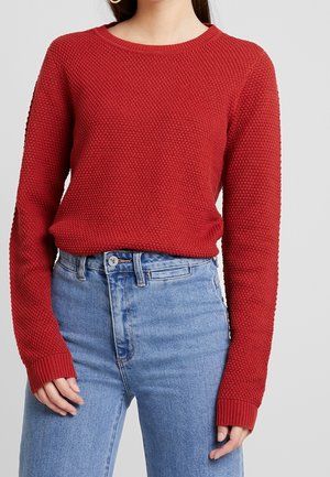 Czerwony sweter z dzianiny z teksturowanym wzorem, okrągłym dekoltem i długimi rękawami, połączony z niebieskimi dżinsami o wysokim stanie.