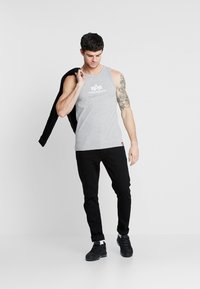 Grå ermeløs tanktopp med en hvit logo, kombinert med sorte bukser og sorte joggesko, som viser en avslappet stil. Modellen holder en jakke.