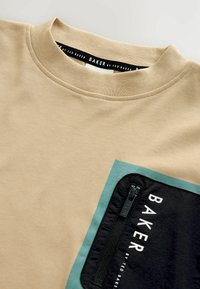Bež pamučna majica s okruglim izrezom. Ima džep s plavim naglaskom, crni zatvarač na mrežici i natpis "BAKER by TED BAKER".