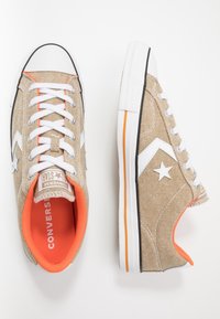 Beżowe płócienne sneakersy z białymi sznurowadłami, z logo gwiazdy, pomarańczowymi akcentami i fakturowaną powierzchnią. Biała gumowa czubek i podeszwa.