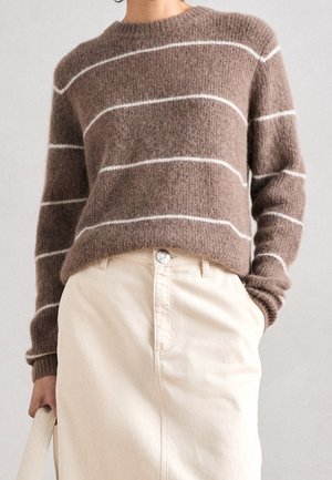 Personne portant un pull marron duveteux avec des rayures blanches horizontales et une jupe crème taille haute, tenant un sac à main crème dans une main.