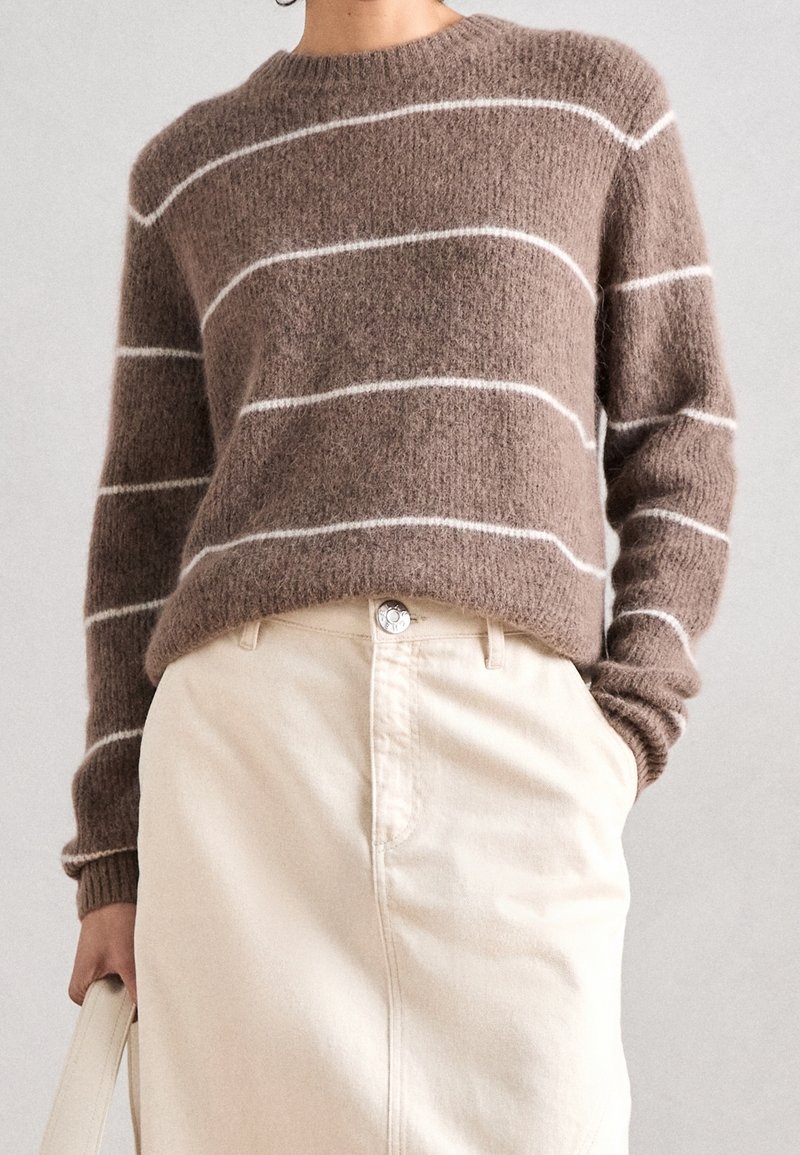 Personne portant un pull marron duveteux avec des rayures blanches horizontales et une jupe crème taille haute, tenant un sac à main crème dans une main.
