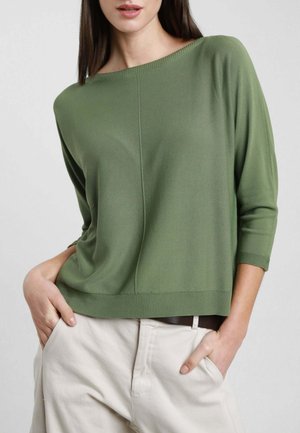 Donna che indossa un maglione verde oliva ampio con maniche a tre quarti e pantaloni beige chiaro, con le mani nelle tasche, davanti a uno sfondo grigio uniforme.