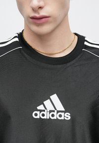 Μαύρο αθλητικό T-shirt με λευκό λογότυπο adidas, με στρογγυλή λαιμόκοψη και τρεις λευκές ρίγες στους ώμους. Ορατό χρυσό κολιέ αλυσίδα.