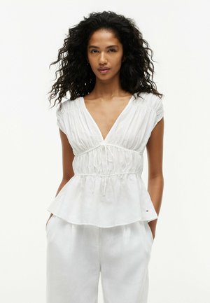 Kobieta o kręconych włosach ubrana w białą bluzkę z krótkim rękawem w serek z talią wciętą oraz białe spodnie, stojąca z rękami w kieszeniach.