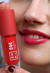 Red lip oil in a clear tube with a matte red cap, displaying the number "244" and logo. The lips are glossy in a similar red shade.