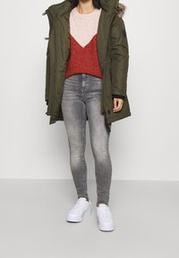 Olivově zelená parka s kapucí podšitou kožešinou, vrstvená přes červený a růžový svetr, v kombinaci se šedými skinny džíny a bílými teniskami.
