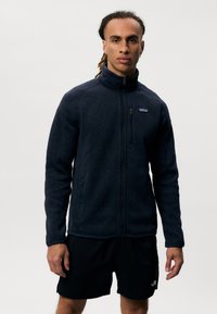 Giacca in pile di colore blu scuro; presenta un alto colletto, zip completa e due tasche laterali. Tessuto morbido con un logo discreto sul petto.