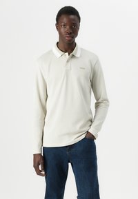 Camisa polo de manga larga color beige con un cuello clásico y tres botones, que presenta un logotipo sutil en el pecho. Se lleva con unos vaqueros azul oscuro.