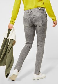 Šedé skinny džíny z denimové látky s vybledlým vzhledem, obsahující zadní kapsy a černou nášivku na pase. Kombinované se zeleným topem a bílými botami.