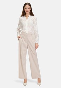 White sheer blouse with floral pattern, buttoned, paired with light beige wide-leg trousers. Model wears pointed white heels.