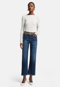 Top bianco a maniche lunghe e jeans in denim a gamba larga blu con dettagli sulle tasche frontali, abbinati a una cintura marrone e tacchi bianchi a punta.